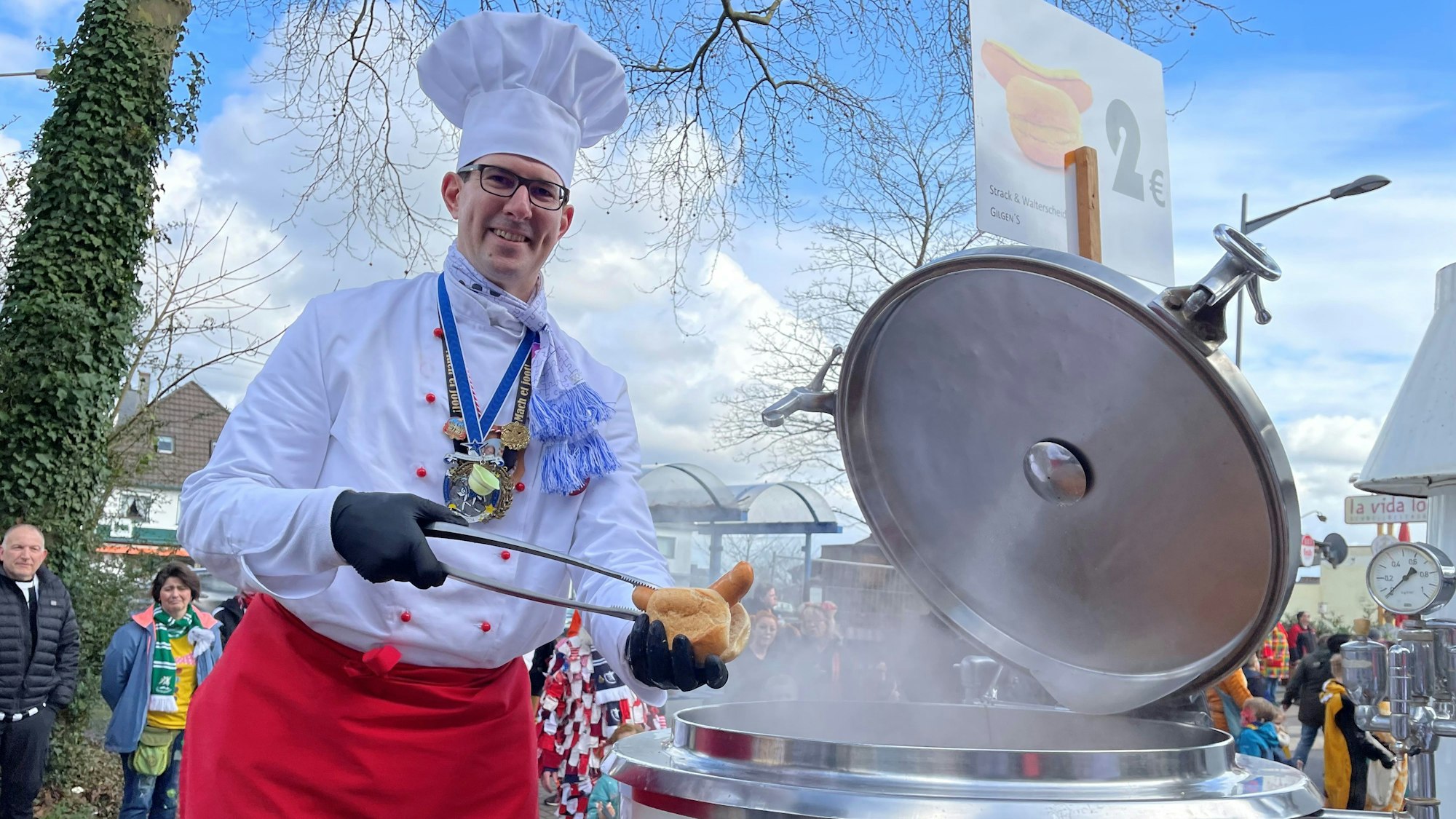 Ein als Koch verkleideter Mann legt mit einer Zange ein Knackwürstchen in ein aufgeschnittenes Brötchen.
