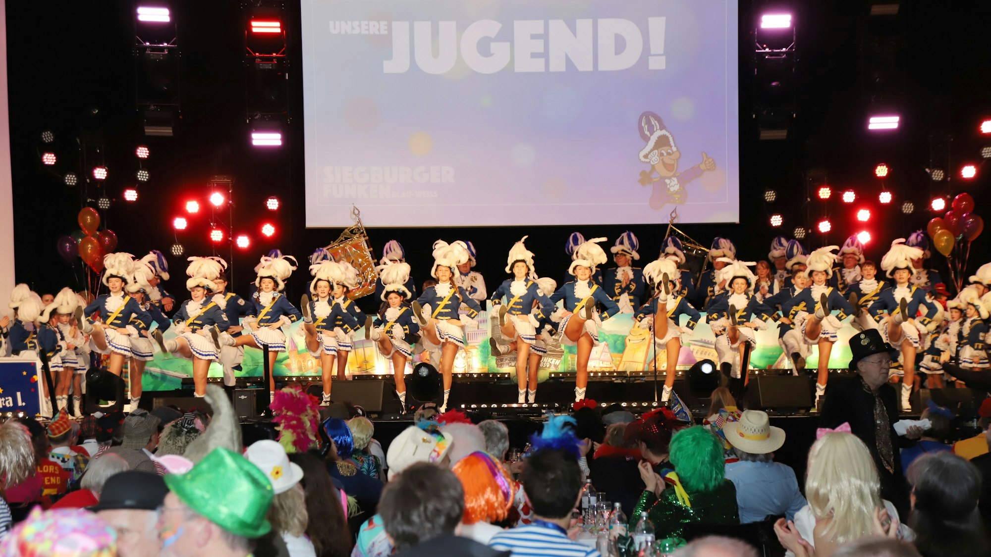 Eine Tanzgarde auf der Bühne eines vollen Saales. Die Besucher sind kostümiert, die Tänzerinnen tragen ihre Gardeuniform.