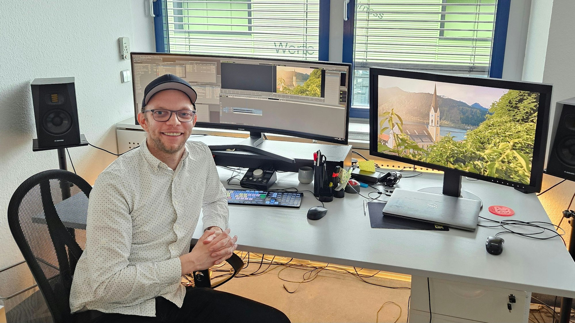 Ein Mann mit Schirmmütze sitzt vor einem Schreibtisch mit zwei großen Bildschirmen und Lautsprecherboxen.