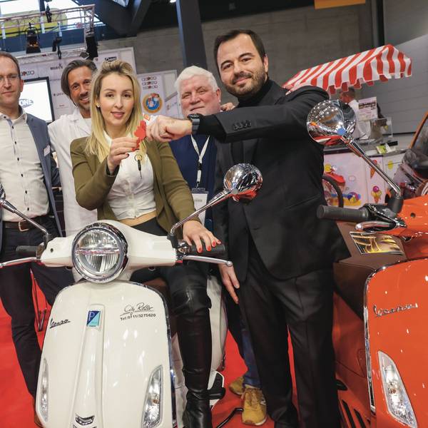 Angelika Szaban sitzt auf einer weißen Vespa. Um sie herum stehen fünf Männer, die mit ihr für ihre Auszeichnung posieren.