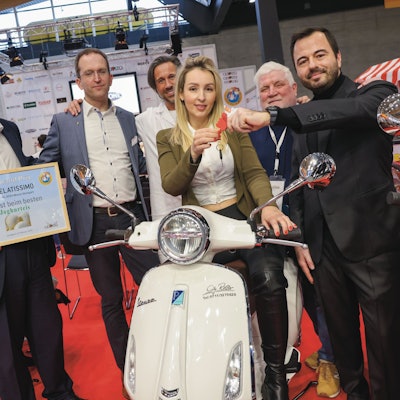 Angelika Szaban sitzt auf einer weißen Vespa. Um sie herum stehen fünf Männer, die mit ihr für ihre Auszeichnung posieren.