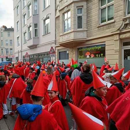 Kinder in roten Kostümen und Spitzhüten stehen als Teil des Veedelszugs auf einer Mülheimer Straße.