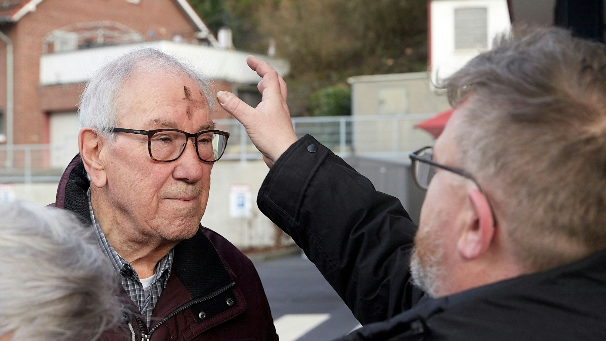 Ein Mann steht auf einem Parkplatz. Ein Pfarrer reibt ihm mit dem Daumen der rechten Hand ein Aschenkreuz auf die Stirn.