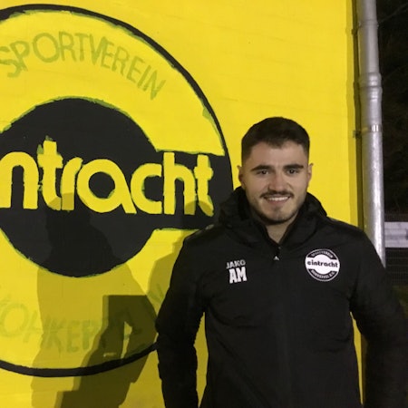 Zu sehen ist ein Fußballer vor einem großen Vereinswappen