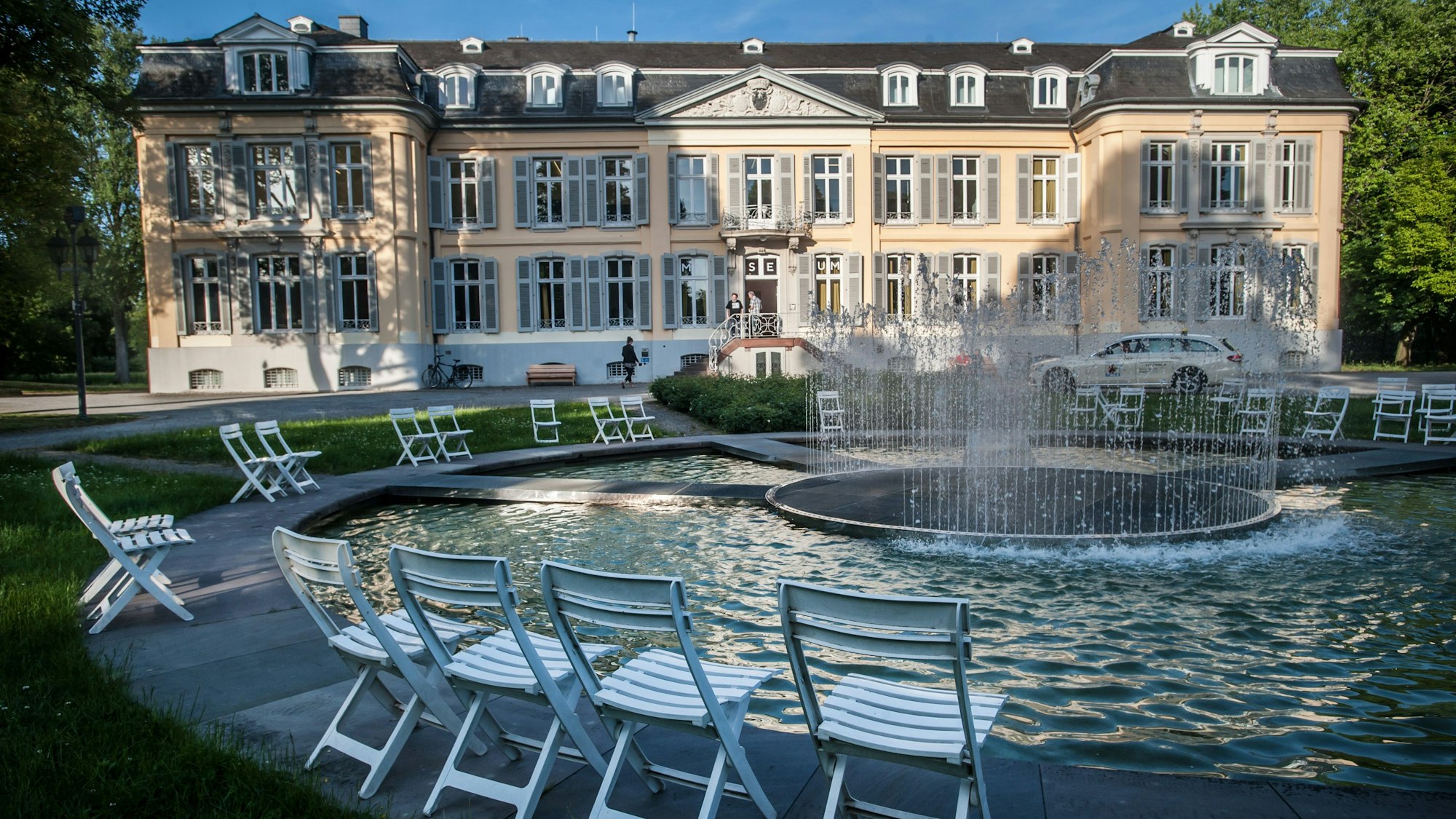 Das Museum Morsbroich von außen. Um einen Springbrunnen stehen Klappstühle verteilt.