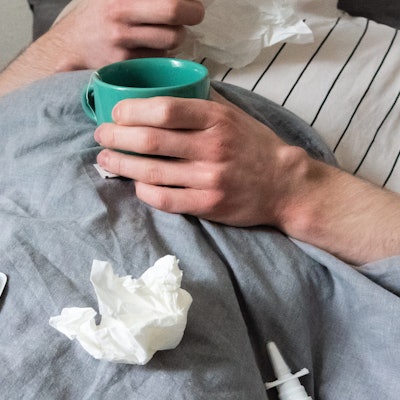 Ein Mann liegt mit Taschentüchern, Teetasse, Nasenspray und Tabletten im Bett (gestellte Szene).
