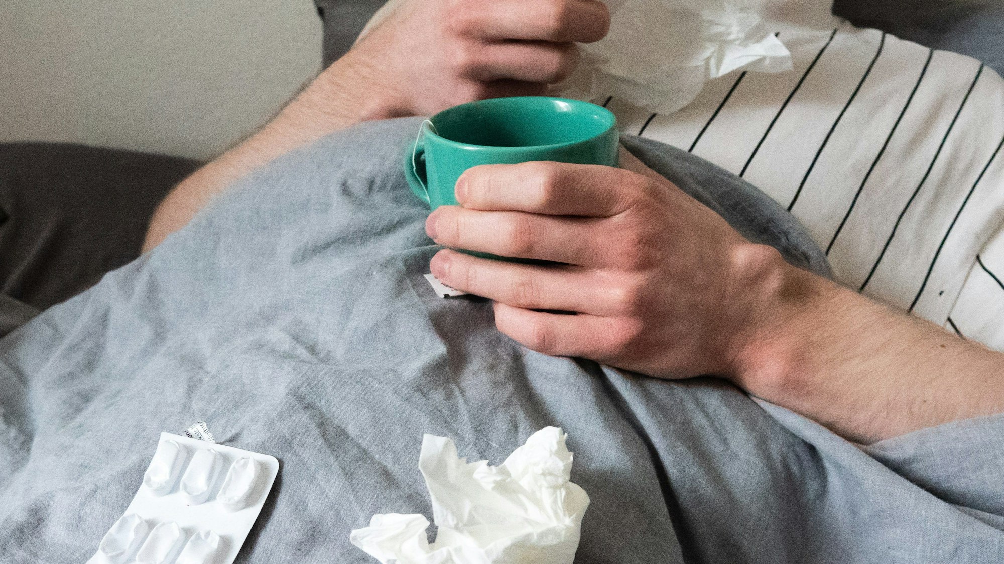Ein Mann liegt mit Taschentüchern, Teetasse, Nasenspray und Tabletten im Bett (gestellte Szene).