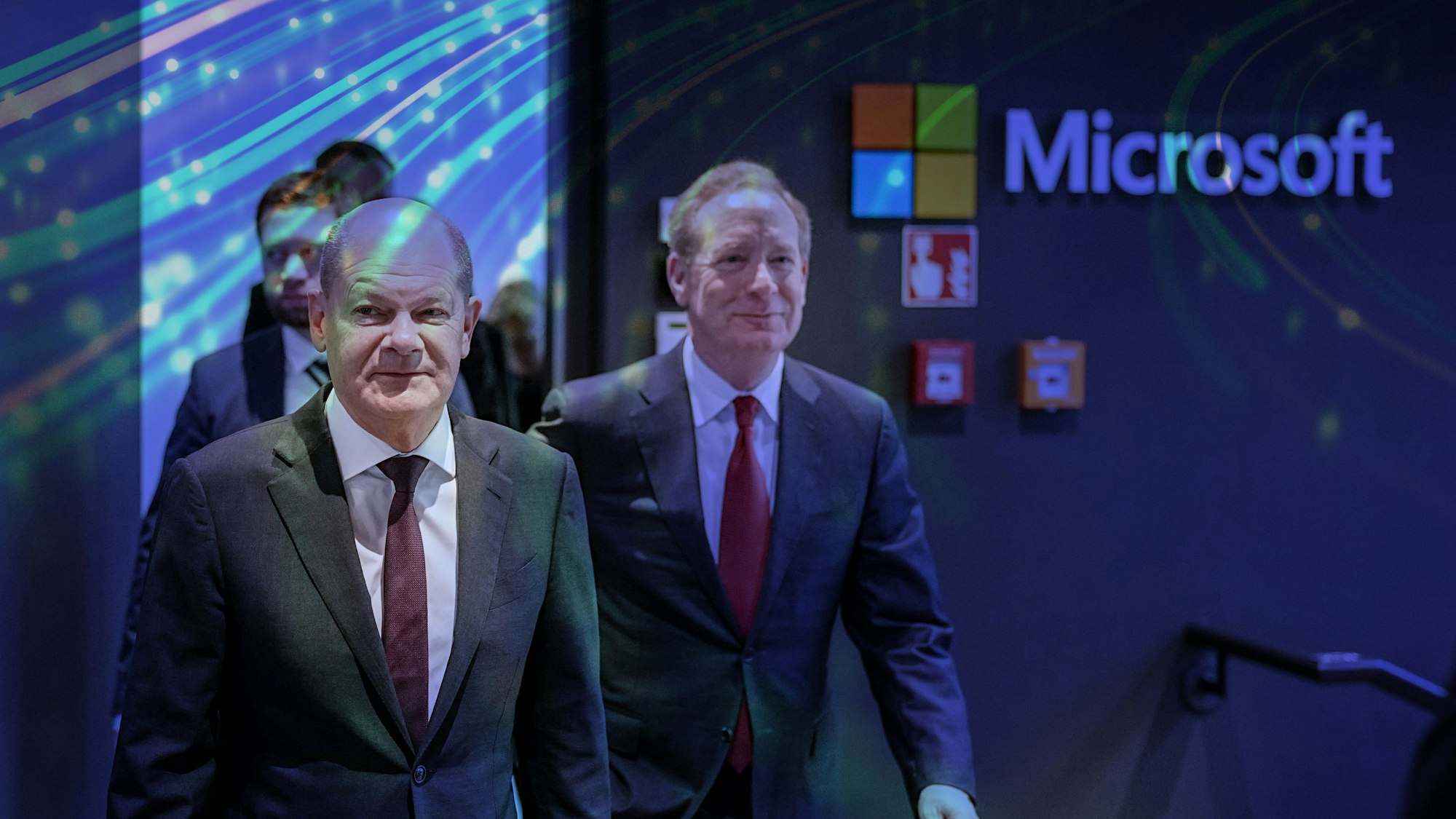 Bundeskanzler Olaf Scholz (SPD) und Microsoft-Präsident Brad Smith auf dem Weg zu einer Pressekonferenz, in der die Investitionspläne des US-Konzerns im Rhein-Erft-Kreis vorgestellt werden. Im Hintergrund ist das Firmenlogo von Microsoft zu sehen, ebenso wie ein Teil einer farbigen Wandpräsentation.