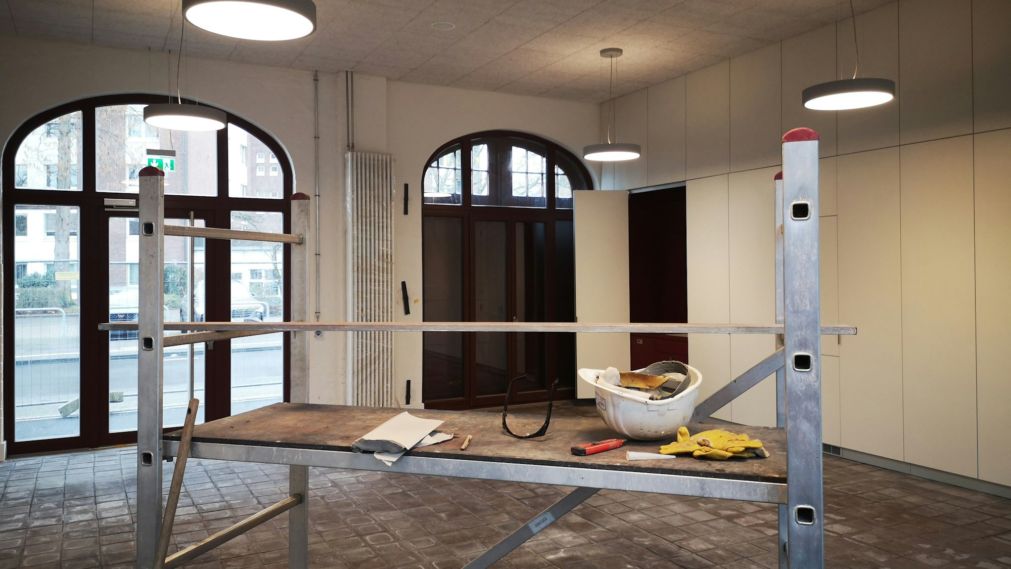 Im Vordergrund ist ein Bauregal mit Bauhelm zu sehen, dahinter die Wagenhalle der Feuerwache mit Rundbogenfenstern.