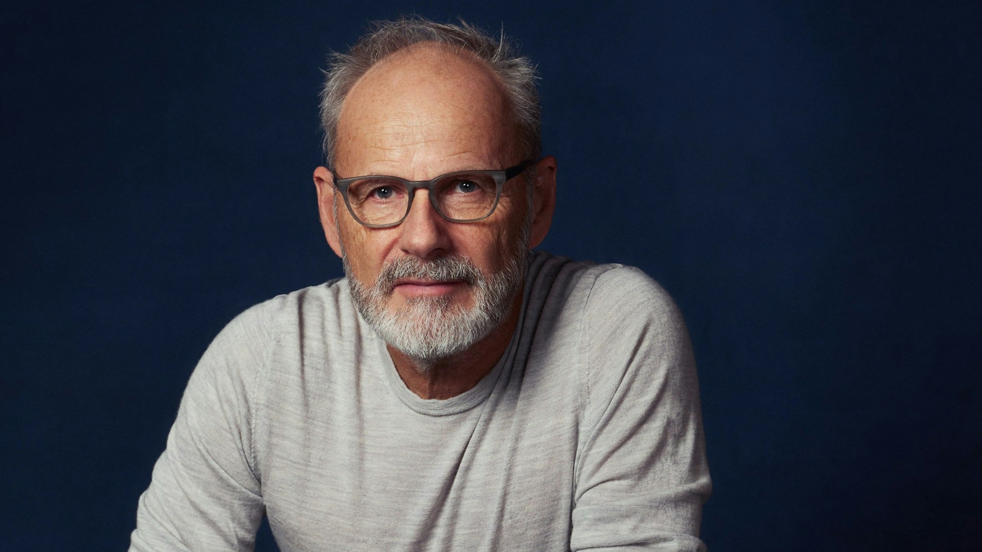 Reinhold Beckmann trägt ein graues Langarm-Shirt, er blickt in die Kamera.
