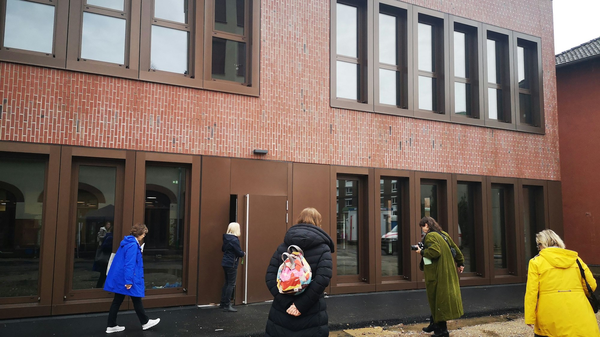 Fünf Personen betreten ein backsteinrotes Gebäude mit vielen Fensterfronten.