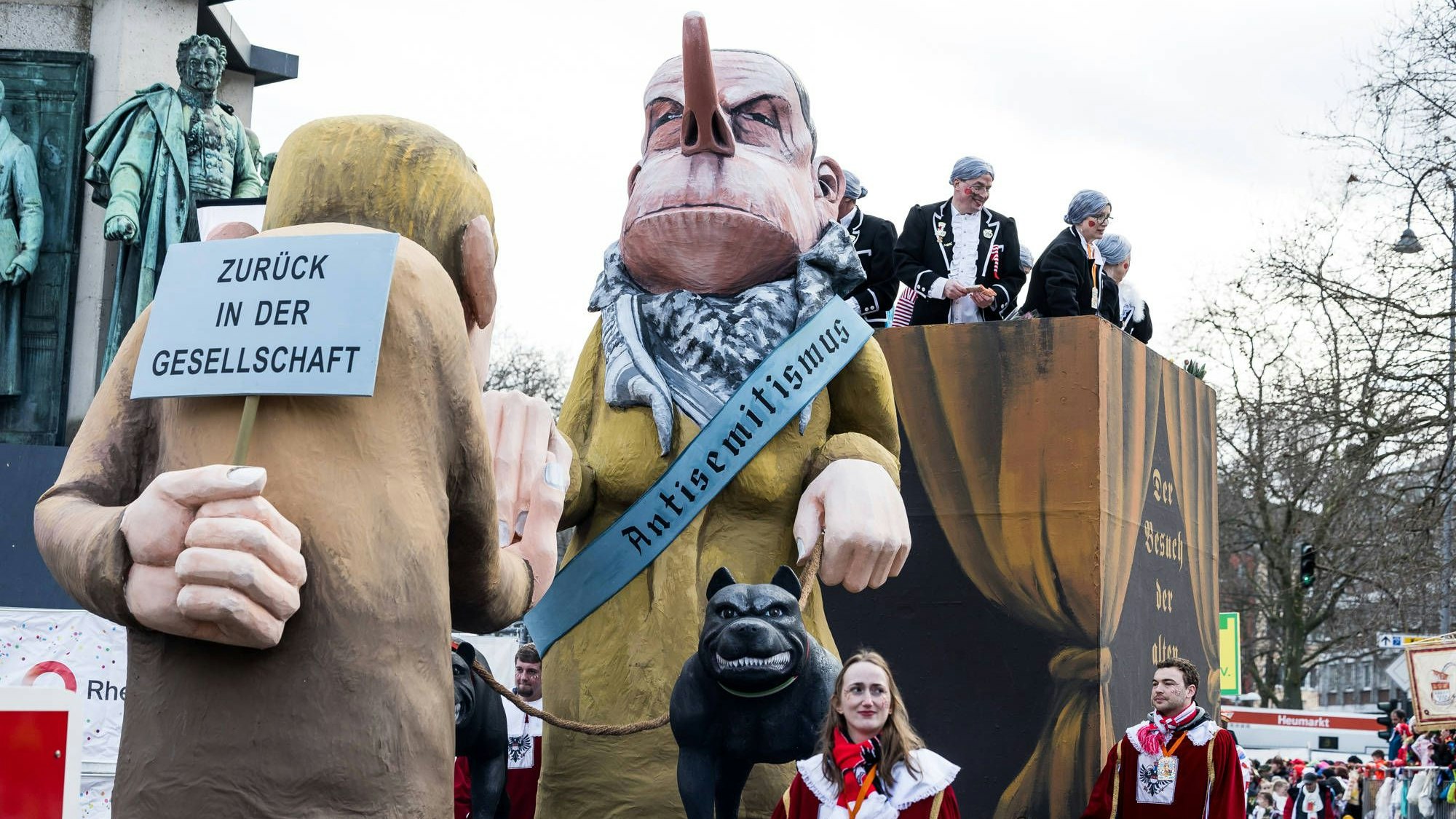 Auf einem Wagen des Kölner Rosenmontagszugs ist eine alte Frau zu sehen, die ein Palästinensertuch trägt sowie eine Schärpe, auf der in gotischer Schrifttype das Wort „Antisemitismus“ steht. Begleitet wird sie von zwei Kettenhunden. Eine blonde Figur in brauner Kleidung steht vor und küsst ihre Hand. Hinter dem Rücken hält die Figur ein Schild mit dem Text „Zurück in der Gesellschaft“, wodurch sie als Symbolgestalt für die neue Rechte erkennbar wird.