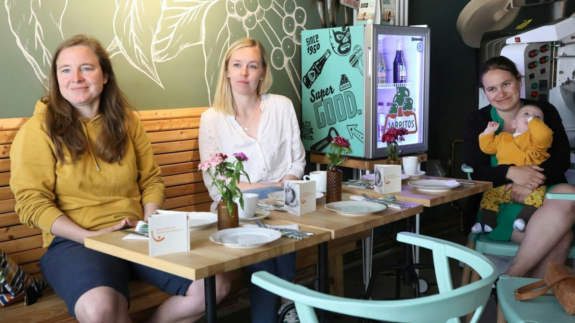 Drei Frauen sitzen an einem Tisch in einem Café, eine hat ein Kleinkind auf dem Arm.
