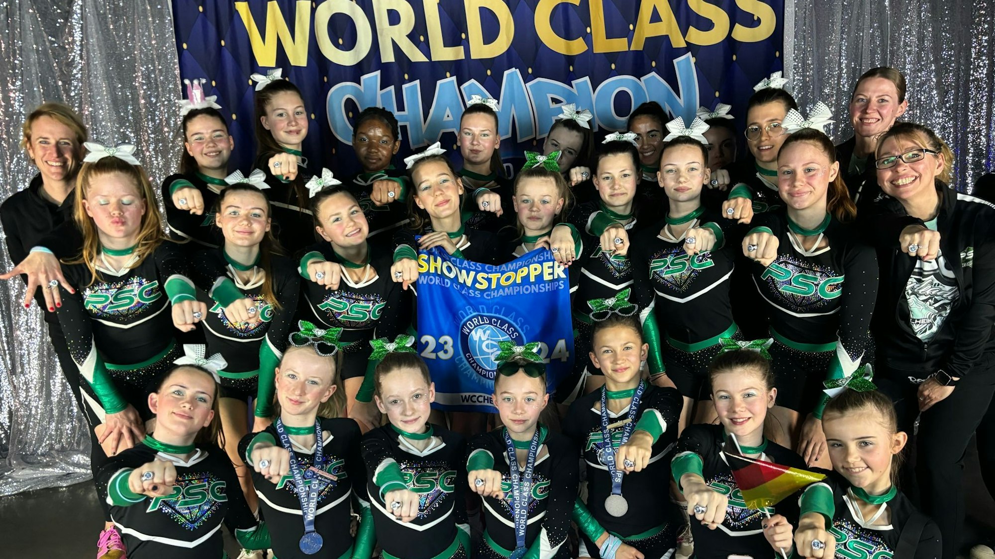 Das Cheerleader-Team und die Trainerinnen strahlen, stolz halten sie ihre Gewinnerringe in die Kamera.