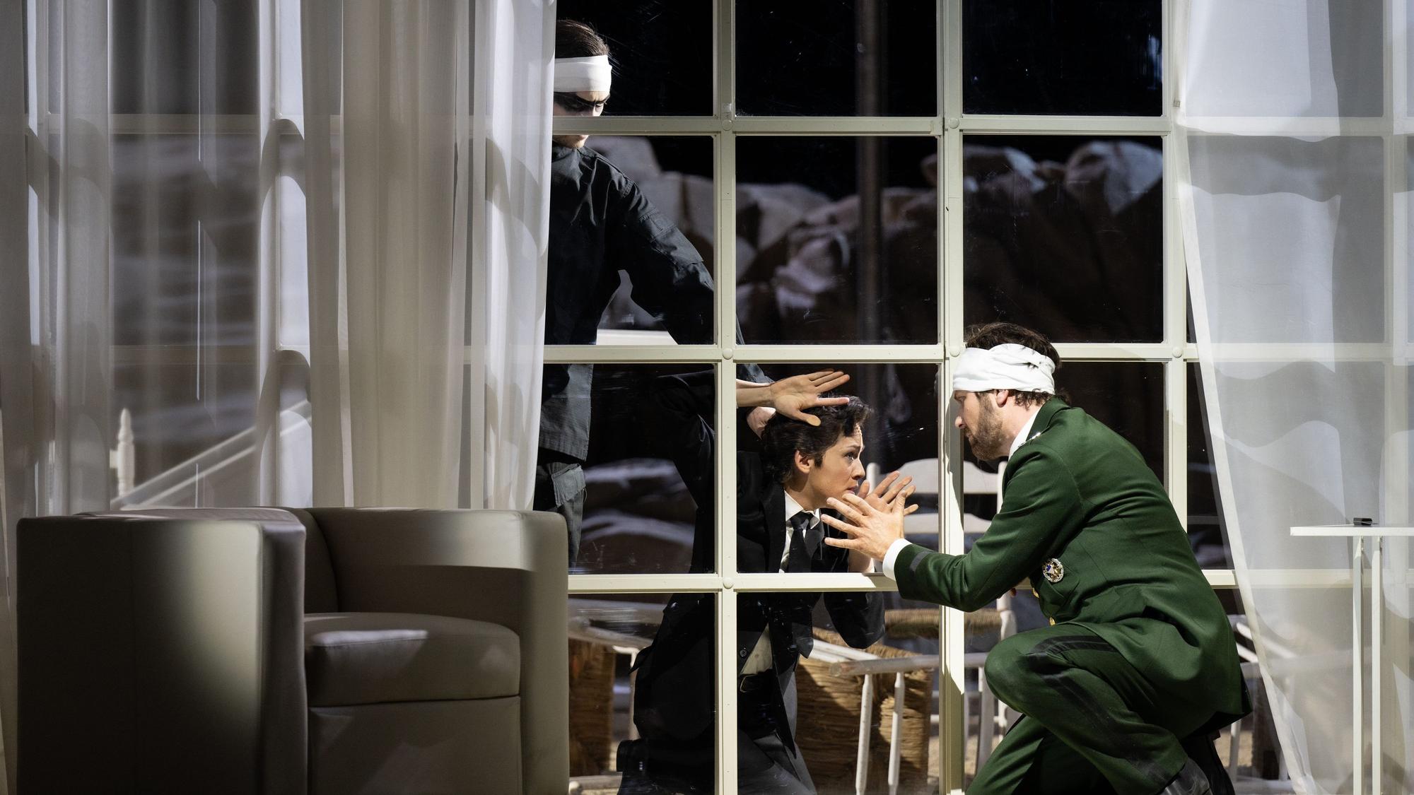 Szene aus der Oper Idomeneo. Ein Mann trägt eine polizeigrüne Uniform und hat einen Verband um den Kopf. Er ist in einem Wohnzimmer und kniet sich vor das Fenster, auf der anderen Seite der Scheibe sitzt eine Frau. Im Hintergrund lauert ein Mann und streckt die Hand nach der Frau aus.