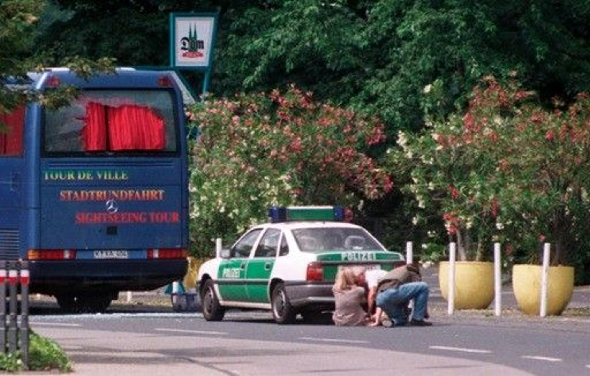 Heck eines Reisebusses und ein Polizeiwagen. Dahinter drei kauernde Menschen