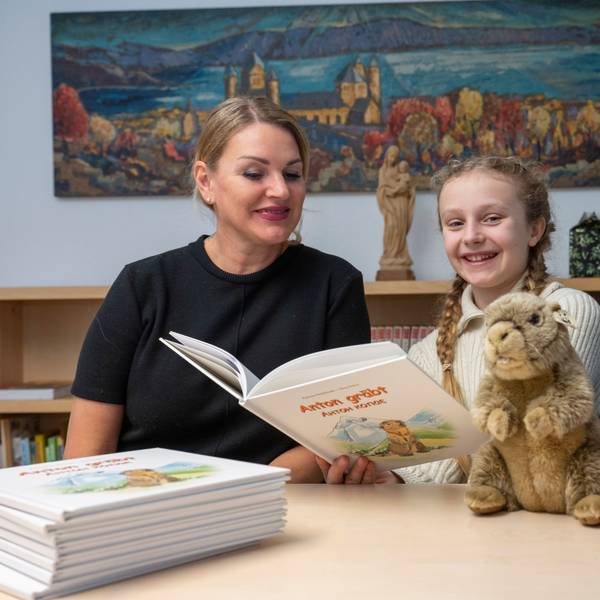 Eine Frau, ein Mädchen und ein Mann sitzen gemeinsam an einem Tisch. Das Mädchen hat ein Buch in der Hand, ein Stapel Bücher und ein Stoffmurmeltier liegen auf dem Tisch.