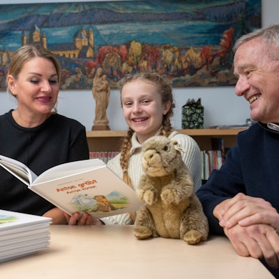 Eine Frau, ein Mädchen und ein Mann sitzen gemeinsam an einem Tisch. Das Mädchen hat ein Buch in der Hand, ein Stapel Bücher und ein Stoffmurmeltier liegen auf dem Tisch.