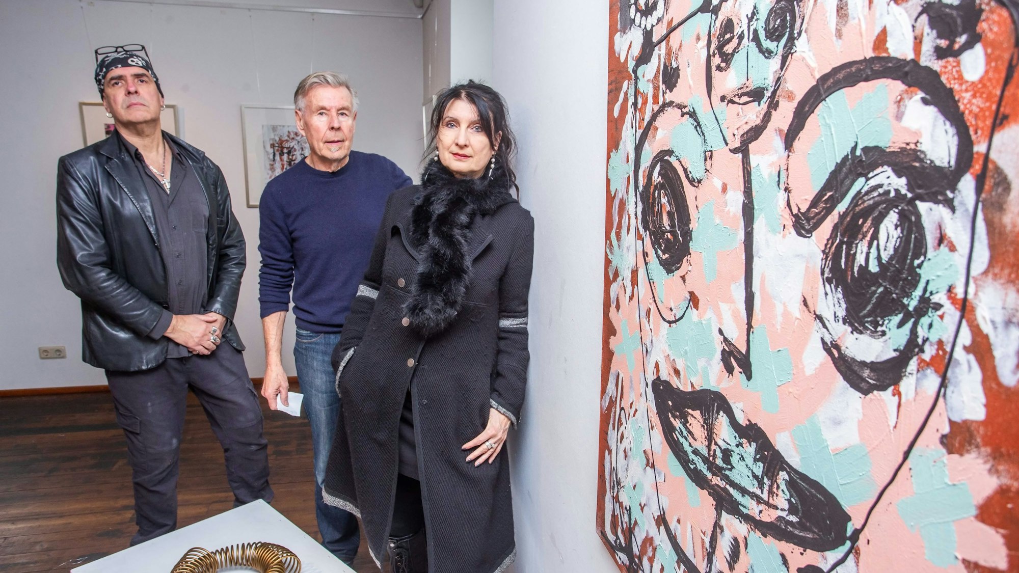 Zwei Männer und eine Frau stehen in einer Ausstellung neben einem großen Bild mit einem gemalten Gesicht.