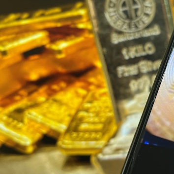 Auf einem Bildschirm sind die Edelmetalle Gold und Silber zu sehen, davor ist ein Smartphone-Bildschirm zu sehen, auf dem das Logo der Kryptowährung Bitcoin zu sehen ist.