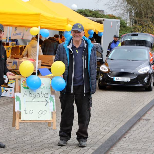 Uli Gürster (l.) von der Humanitären Hilfe Bergisch Gladbach und Norbert Kuhl (r.) von der Humanitären Hilfe Overath halten ein Schild mit der Aufschrift „Abgabe to go“ am Spenden-Drive-In ihrer Vereine im Frühjahr 2023.