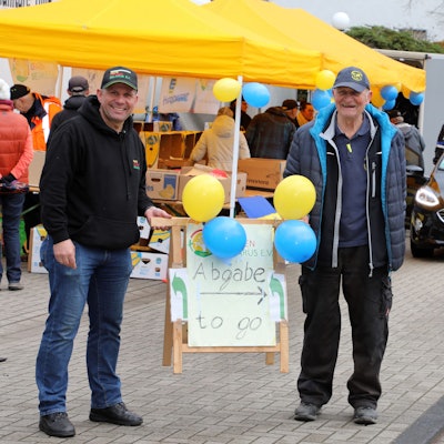 Uli Gürster (l.) von der Humanitären Hilfe Bergisch Gladbach und Norbert Kuhl (r.) von der Humanitären Hilfe Overath halten ein Schild mit der Aufschrift „Abgabe to go“ am Spenden-Drive-In ihrer Vereine im Frühjahr 2023.
