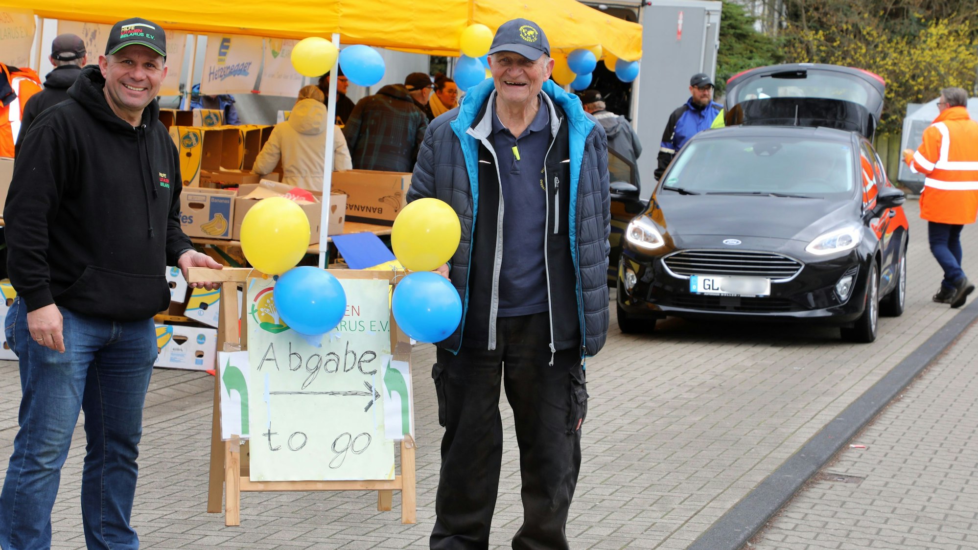 Uli Gürster (l.) von der Humanitären Hilfe Bergisch Gladbach und Norbert Kuhl (r.) von der Humanitären Hilfe Overath halten ein Schild mit der Aufschrift „Abgabe to go“ am Spenden-Drive-In ihrer Vereine im Frühjahr 2023.