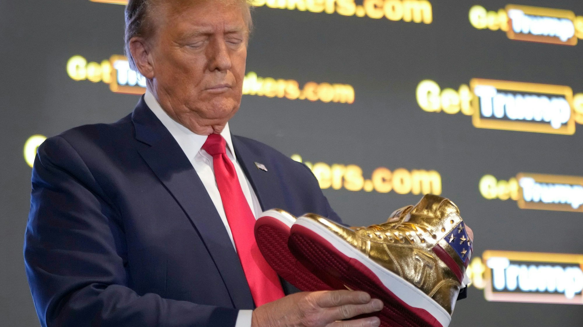 Donald Trump hält goldene Trump-Sneakers  in Händen.