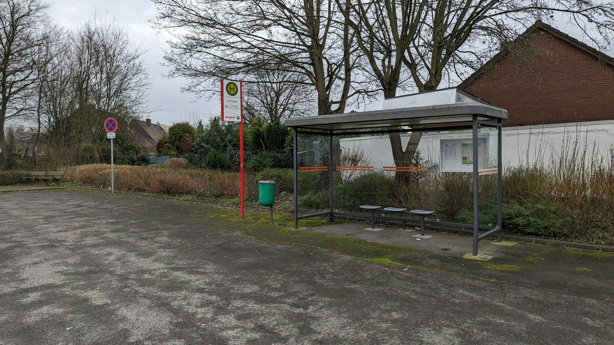 Die Bushaltestelle Im Eisholz steht mitten auf einem Schotterplatz, drumherum ist nicht viel. Im Hintergrund ist die Rückseite eines Hauses zu sehen.