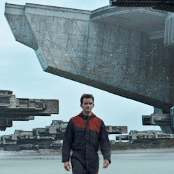 Im Vordergrund des Bildes geht ein Mann in einem Overall am Strand entlangspazieren, im Hintergrund schweben viele riesige Raumschiffe über dem Boden.