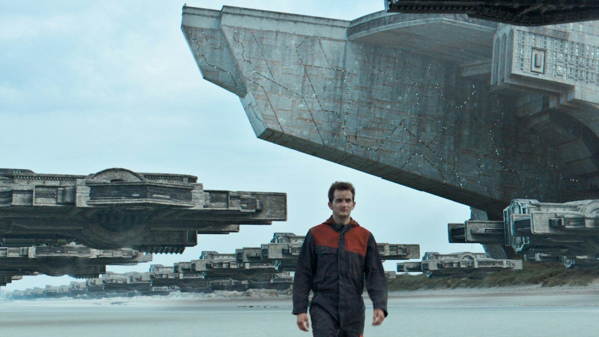 Im Vordergrund des Bildes geht ein Mann in einem Overall am Strand entlangspazieren, im Hintergrund schweben viele riesige Raumschiffe über dem Boden.