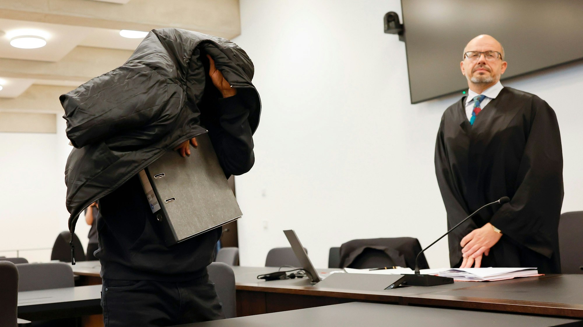 Der angeklagte Ex-Mitarbeiter eines Sicherheitsdienstes (l) bedeckt zu Prozessbeginn seinen Kopf mit einer Jacke. Dahinter steht sein Verteidiger Inigo Schmitt-Reinholtz.