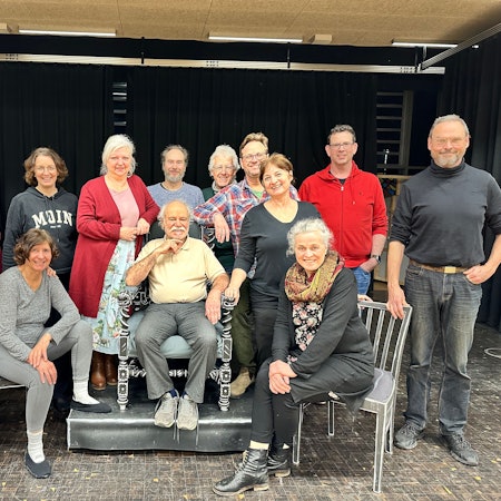 Eine Theatergruppe steht und sitzt auf einer Bühne.