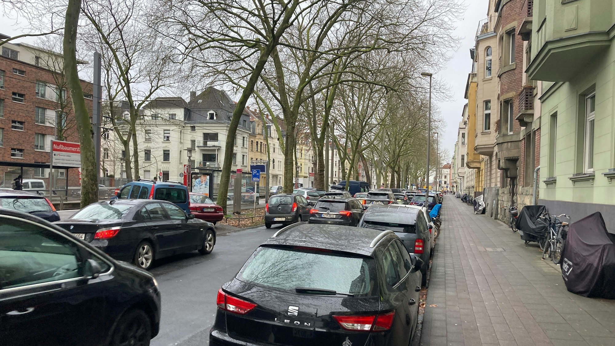 Autos fahren auf zwei Spuren in Richtung Westen über den Ehrenfeldgürtel. Am Straßenrand parken Autos.