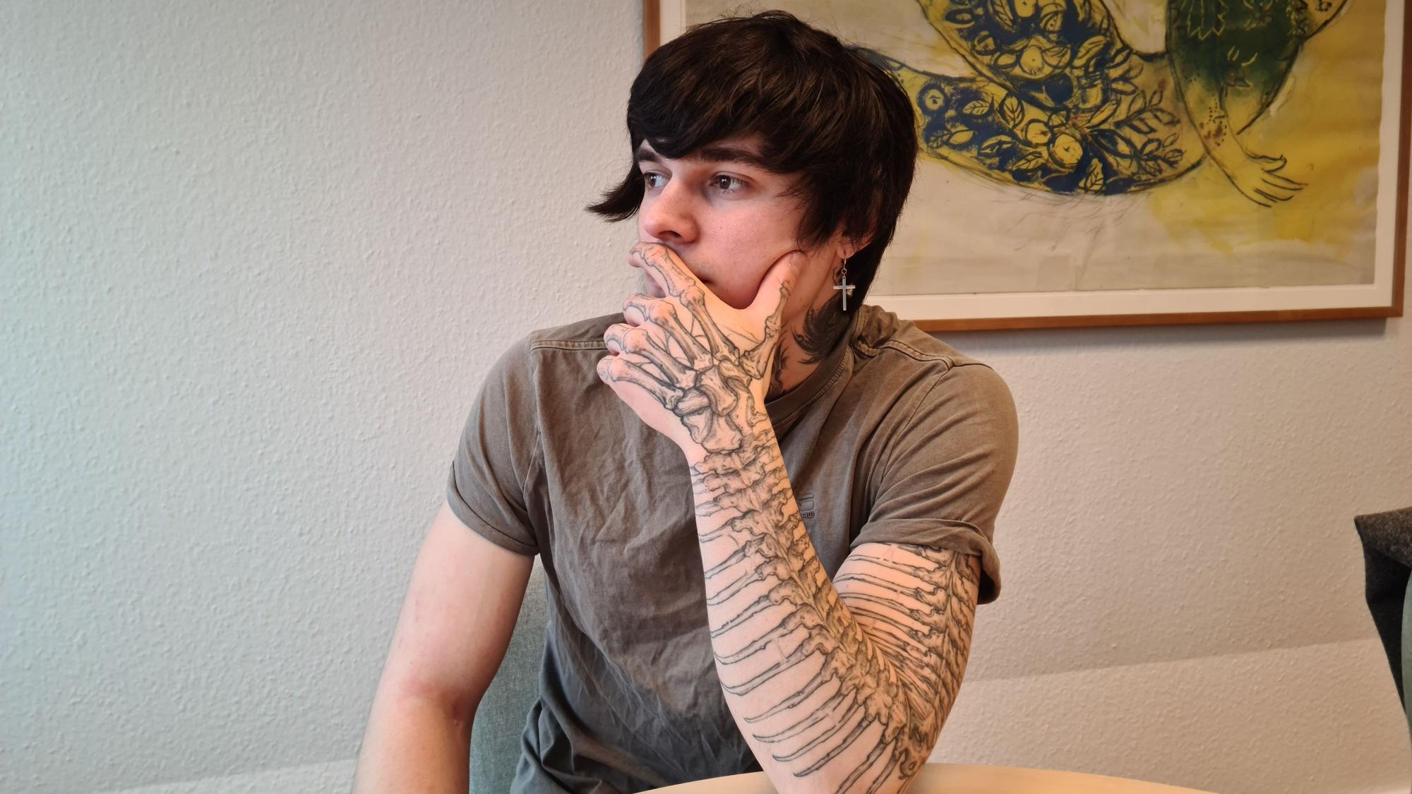 Ein junger Mann sitzt an einem Tisch. Sein aufgestützter Arm ist von oben bis unten tätowiert.