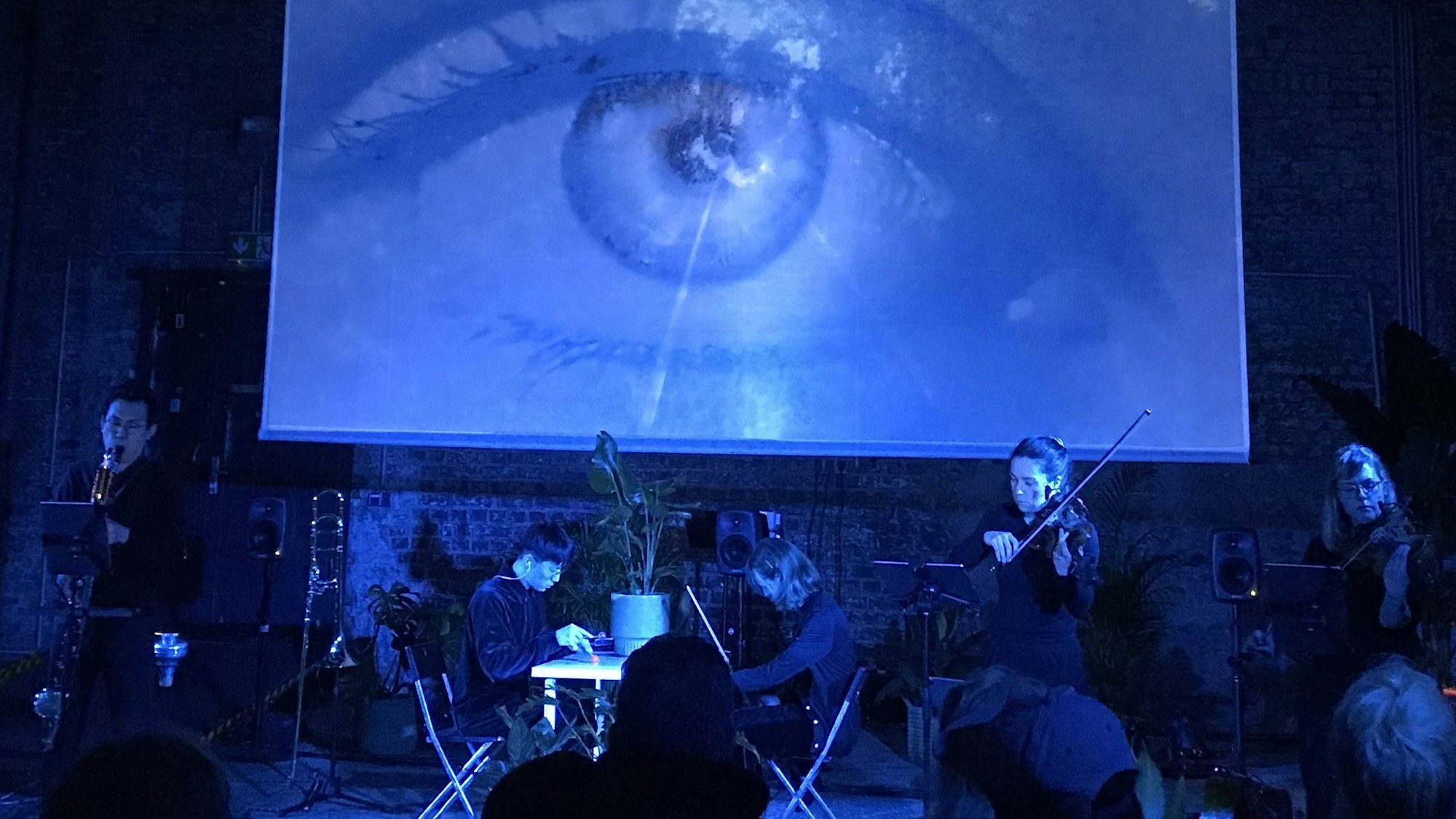 Die Bühne ist dunkel und bläulich erleuchtet. Mehrere Musiker spielen ihre Instrumente, über ihnen wird auf einer großen Leinwand ein riesiges Auge projiziert.
