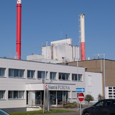 Das Bild zeigt den Eingang zum Verwaltungstrakt der Fabrik, im Hintergrund zwei Schornsteine.