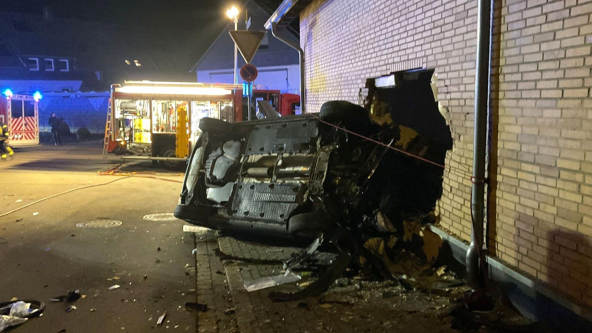 Bei einem Verkehrsunfall ist ein Pkw in eine Hauswand gefahren. Das Fahrzeug liegt auf der Seite, in der Hauswand befindet sich ein großes Loch. Im Hintergrund ist ein Einsatzfahrzeug der Feuerwehr zu sehen.