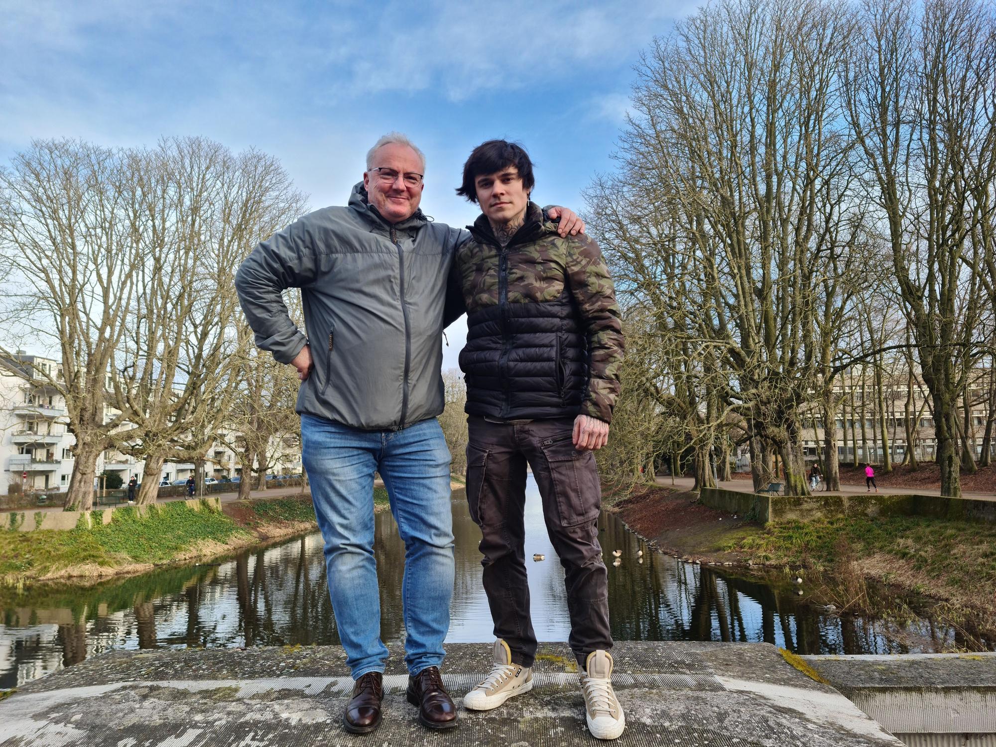 Ein älterer und ein jüngerer Mann stehen nebeneinander auf einer Mauer über dem Lindenthaler Kanal.