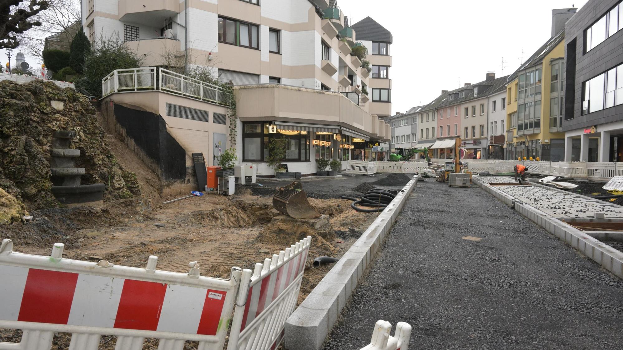 Die Fahrbahn ist bereits mit Asphalt ausgekoffert, rechts und links sind Randsteine verlegt. Ein Teil des Fußwegs rechts ist schon mit dem neuen Pflaster ausgestattet. Links vor dem Emilienbrunnen ist noch Erde. Im Vordergrund steht eine rot-weiße Bake.