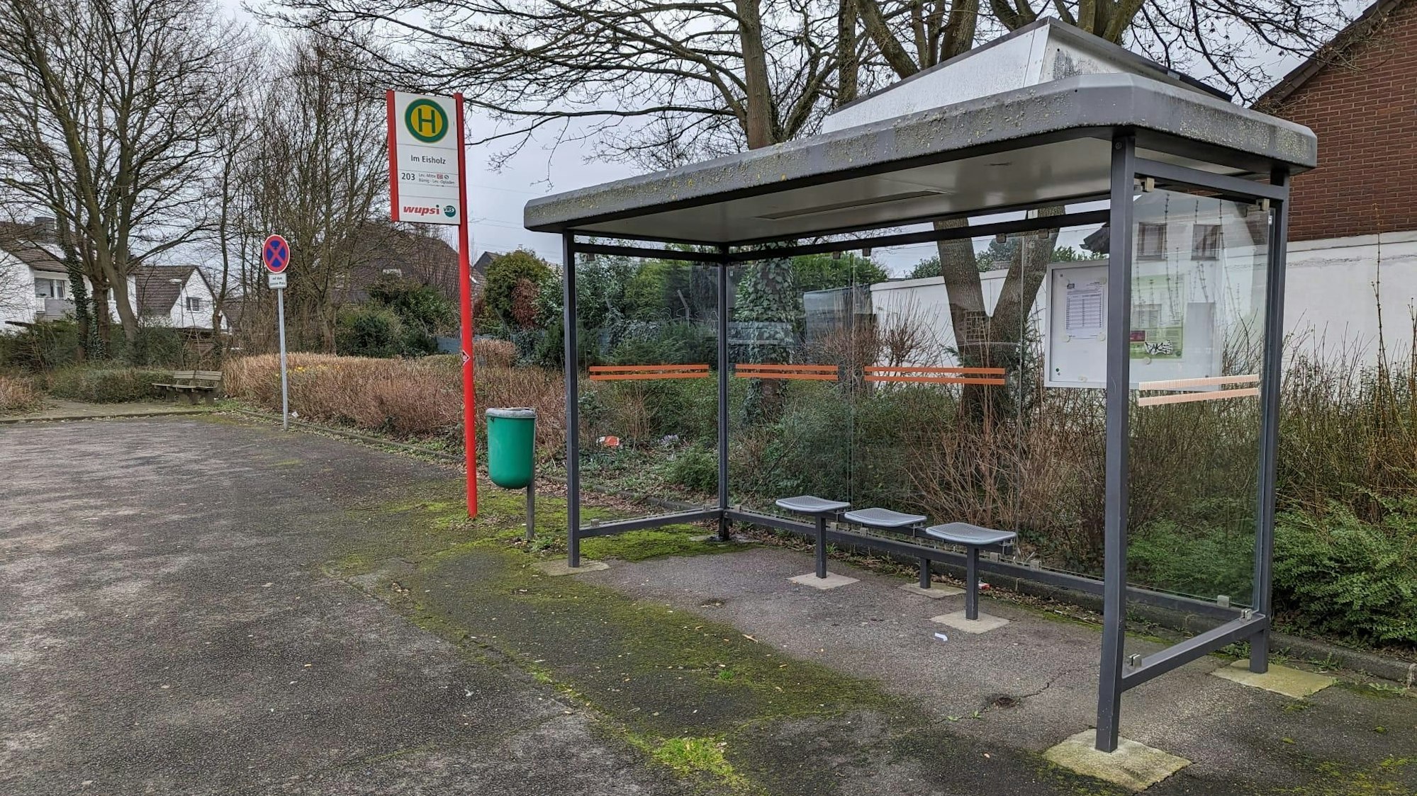 Die Bushaltestelle Im Eisholz steht mitten auf einem Schotterplatz, drumherum ist nicht viel. Im Hintergrund ist die Rückseite eines Hauses zu sehen.