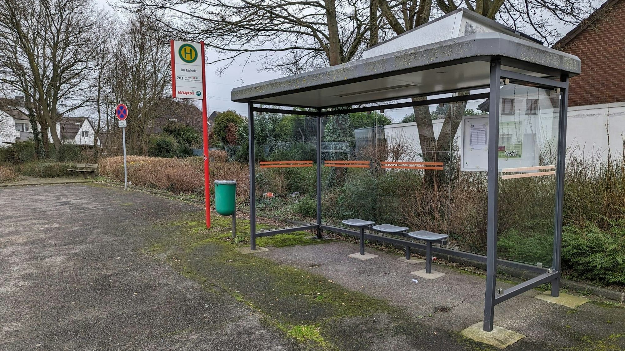 Die Bushaltestelle Im Eisholz steht mitten auf einem Schotterplatz, drumherum ist nicht viel. Im Hintergrund ist die Rückseite eines Hauses zu sehen.