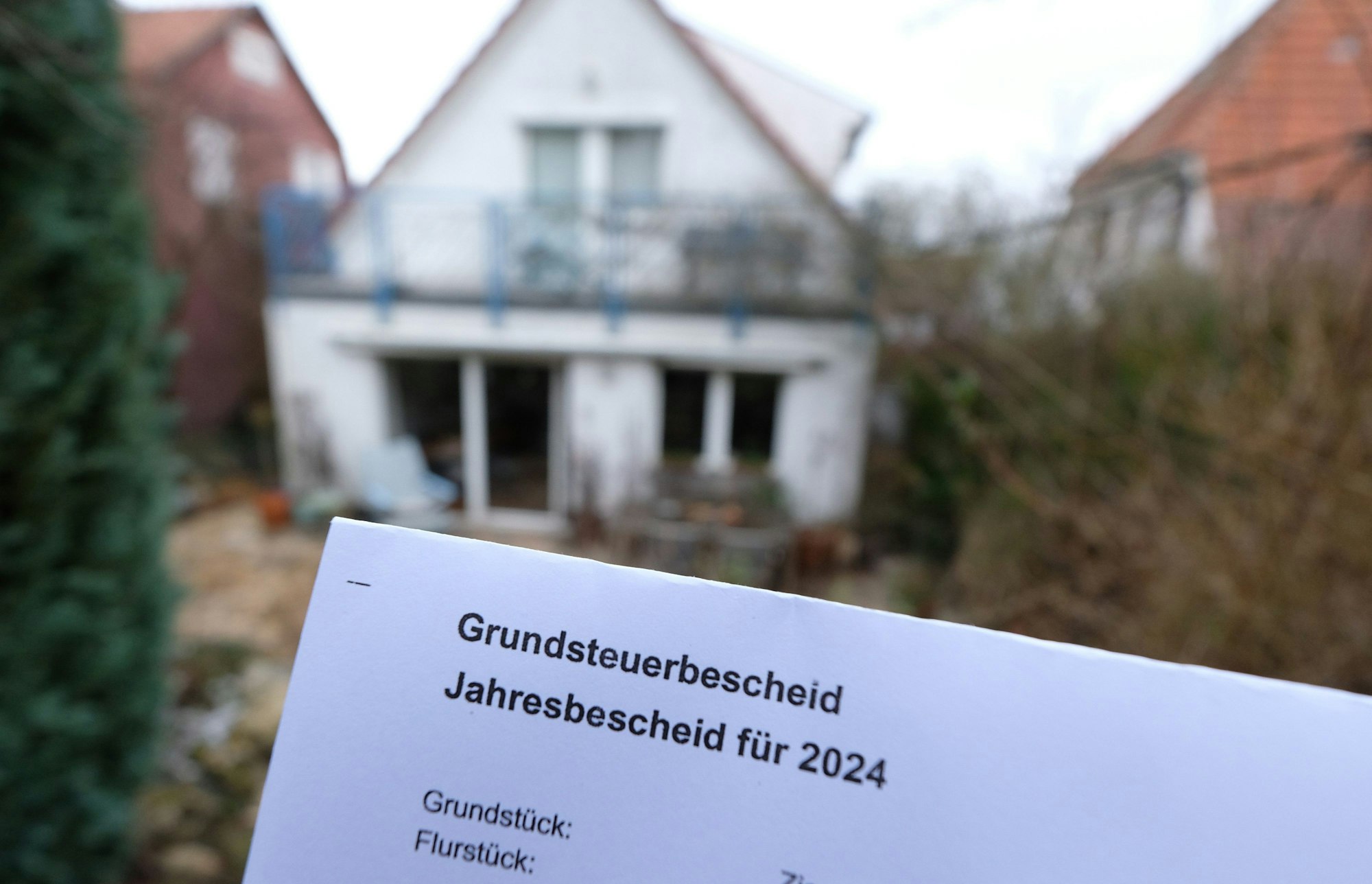 Ein Grundsteuerbescheid für 2024 wird vor einem Privatgrundstück hochgehalten. Im Bildhintergrund sind unscharf mehrere Einfamilienhäuser zu sehen.