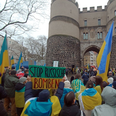 Zu sehen sind Demonstranten, die auf dem Kölner Rudolfplatz stehen und blau-gelbe Fahnen und Kleidung tragen.