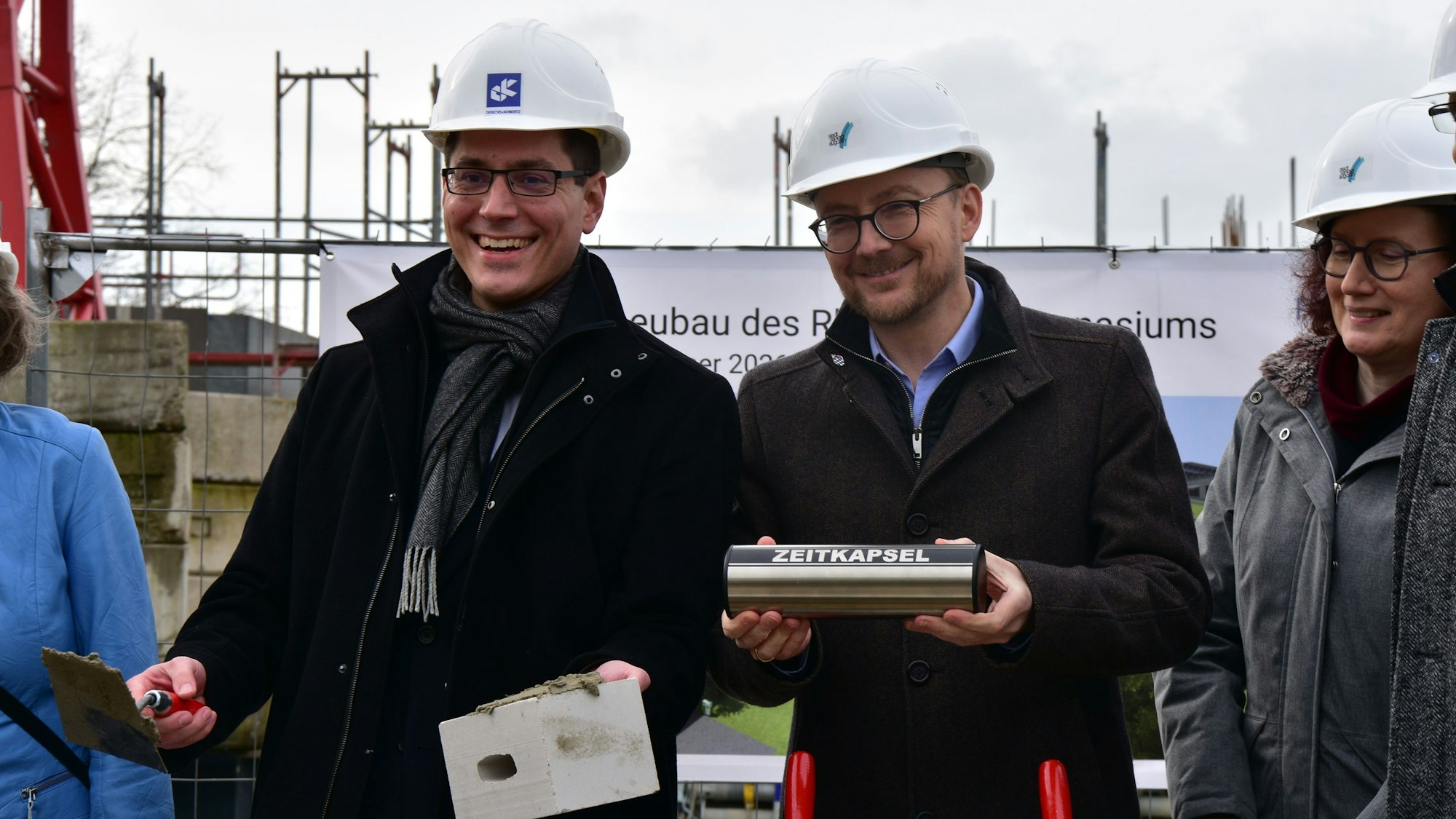 Bürgermeister und Beigeordneter halten Stein und Mörtel, sowie Zeitkapsel in der Hand.