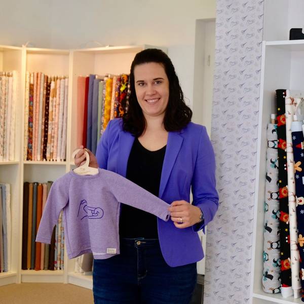 Eine Frau mit dunklem schulterlangem Haar steht vor einem Regal mit Stoffen. Sie zeigt einen Kinderpulli auf einem Bügel.