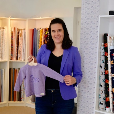 Eine Frau mit dunklem schulterlangem Haar steht vor einem Regal mit Stoffen. Sie zeigt einen Kinderpulli auf einem Bügel.