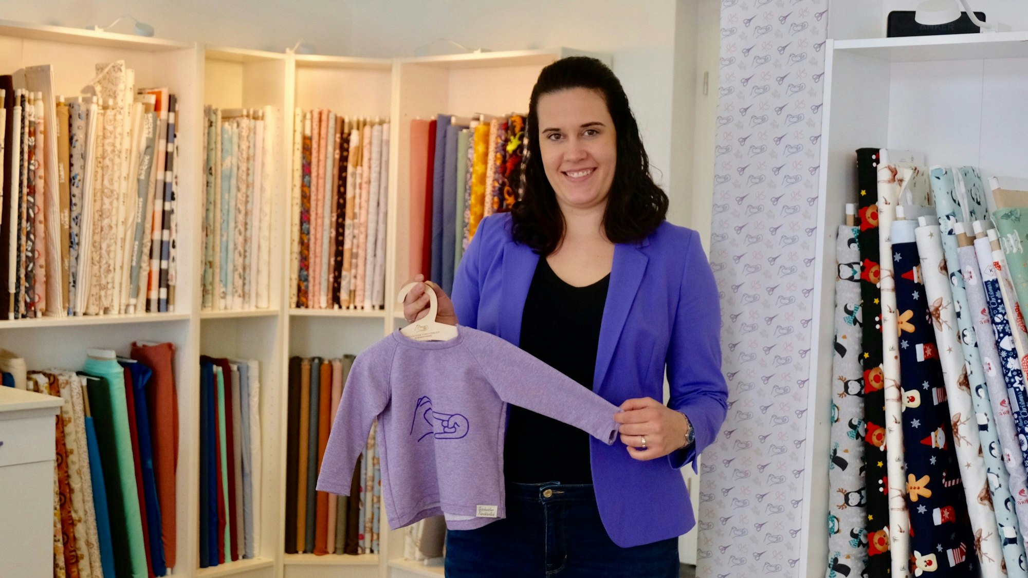 Eine Frau mit dunklem schulterlangem Haar steht vor einem Regal mit Stoffen. Sie zeigt einen Kinderpulli auf einem Bügel.
