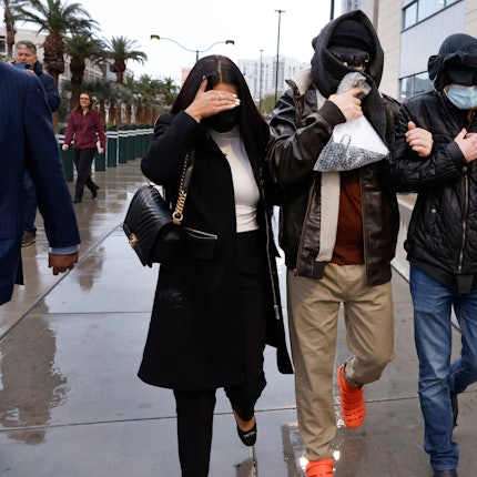 Drei eingehakte Personen gehen maskiert über eine verregnete Straße - im Hintergrund stehen Palmen und Gebäude.