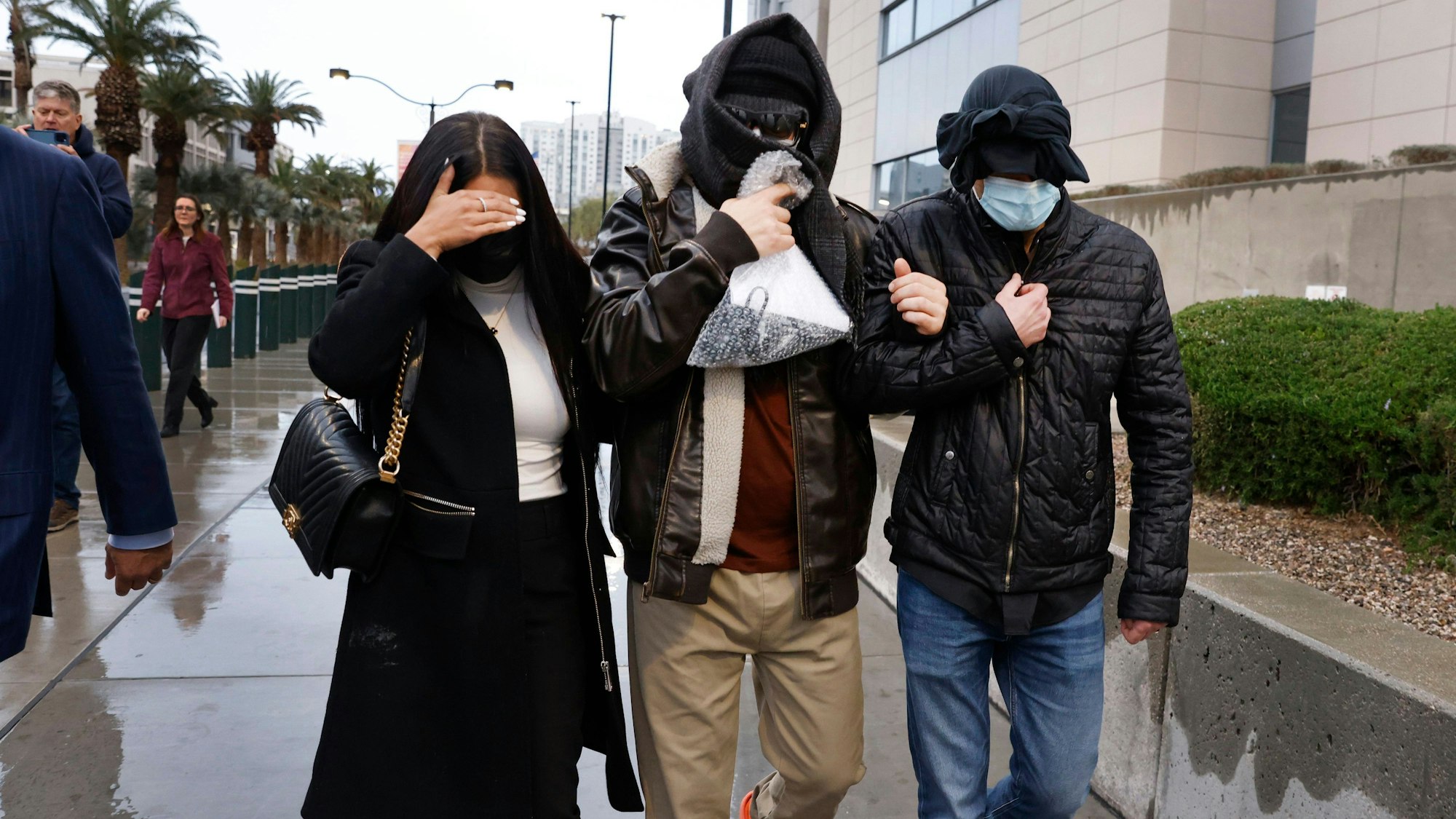 Drei eingehakte Personen gehen maskiert über eine verregnete Straße - im Hintergrund stehen Palmen und Gebäude.